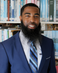 DeVonte Bess Professional headshot of DeVonte Bess smiling and looking directly at the camera. He is wearing business attire and is photographed in front of a bookshelf filled with books