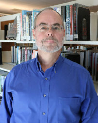 Professional headshot of Grady Miller smiling and looking directly at the camera. He is wearing business attire and is photographed in front of a bookshelf filled with books