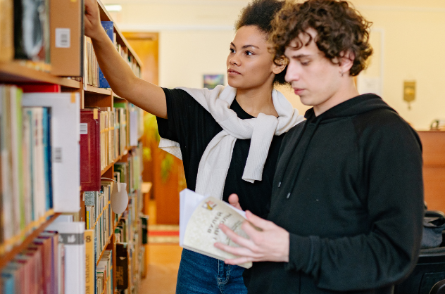 Two teens browse books in a library. One reaches for a book on a shelf while the other flips through a book.