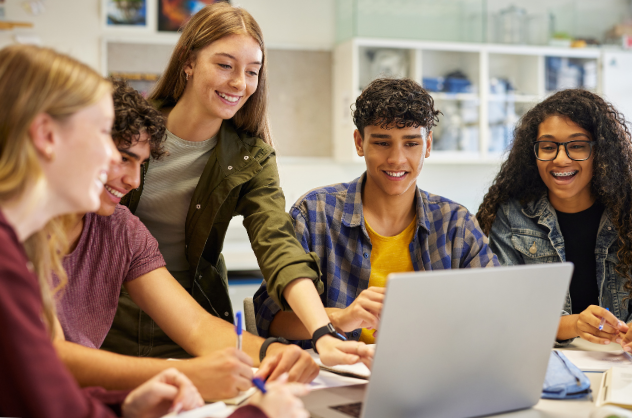 Teens sit around a table studying on a laptop while a standing student points to the screen, and the group smiles.