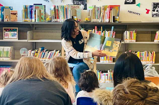 Storytime leader smiling while reading a book to children and their caregivers.